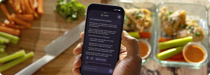 A hand holding a smartphone showing the Stelo app's Coach feature answering the question "Am I trending better than yesterday?"