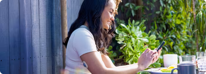 A woman sitting at an outdoor café smiling while browsing her smartphone.