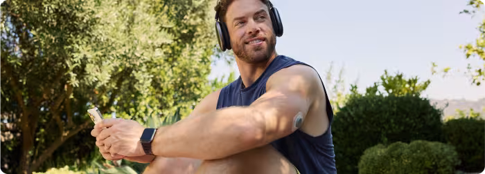 A man sitting outside, wearing a CGM on his upper arm, dressed in workout clothes and sporting headphones.